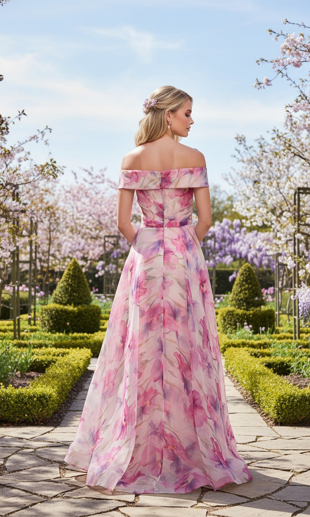 Wearing the La Femme 33710 Long Prom Dress, a woman in a pink floral off-the-shoulder gown with a chiffon A-line skirt stands on a garden path, surrounded by manicured hedges and blooming trees, facing away.